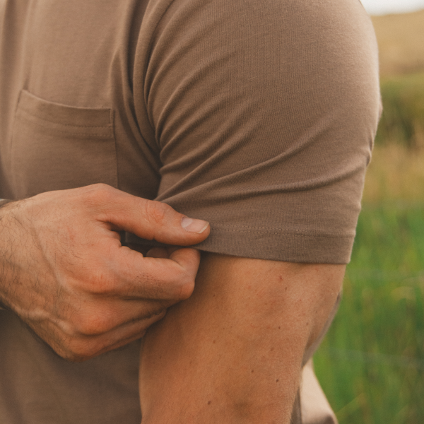 Fingers pinching the sleeve of a tan cotton-modal Shinesty tee, the fabric stretching between them