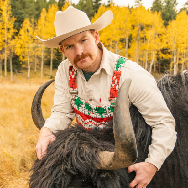 Mustached cowboy crouched beside a yak, front chest pocket of his red and green pajamaralls in view
